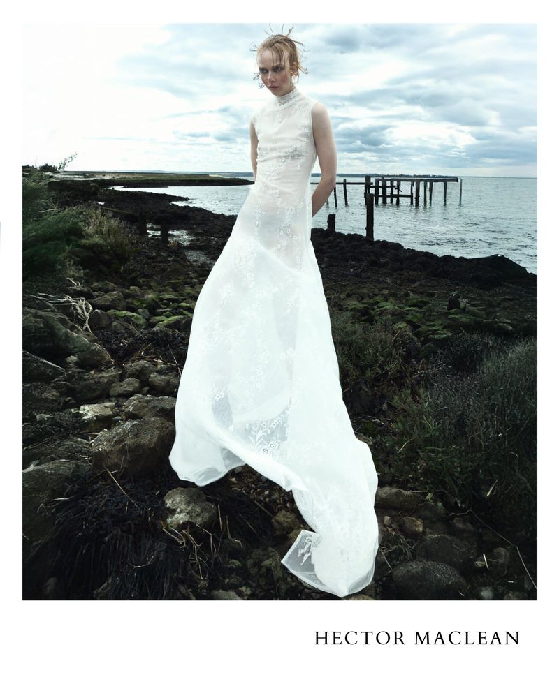 Model Lily Nova standing on a rocky shoreline wearing a sleeveless white lace gown for Hector Maclean.