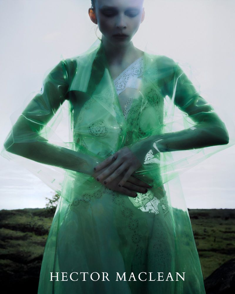 Model Lily Nova wears a translucent green coat over a white lace dress in a moody outdoor landscape for Hector Maclean.