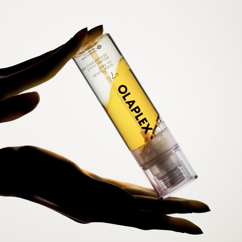 Silhouetted hands holding a clear Olaplex No 7 Shine Serum Oil Mist bottle with golden liquid against a bright white background