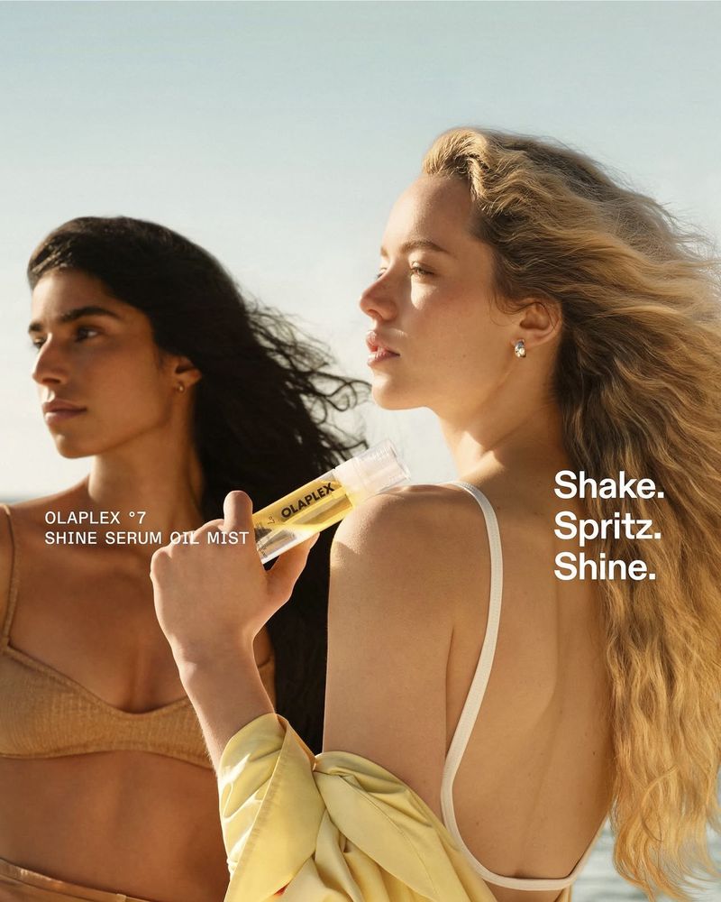 Two women outdoors under bright sunlight, one holding a bottle of Olaplex Shine Serum Oil Mist against her shoulder.