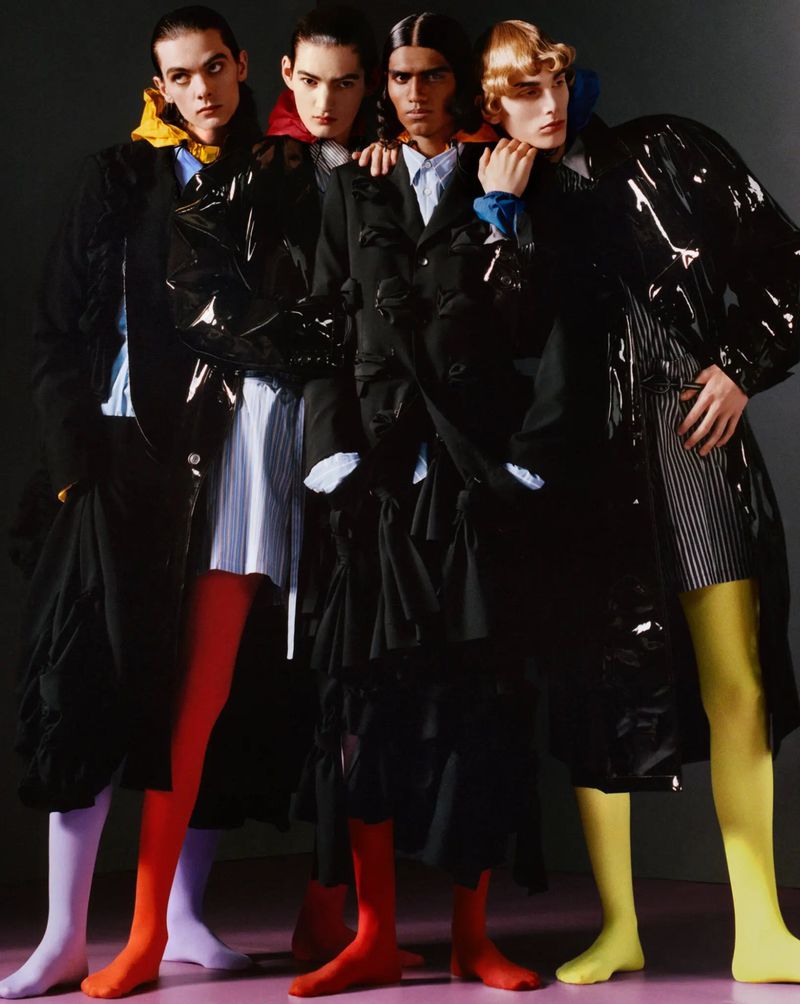 Four male models posing in avant-garde black tailored coats with vibrant colored tights against a dark studio backdrop.