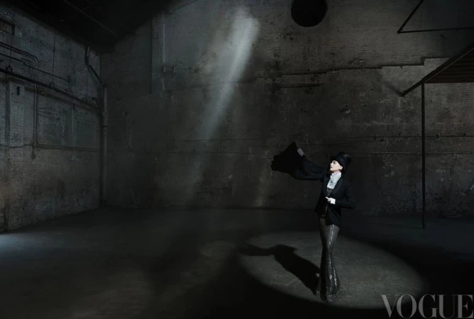 Anne Hathaway standing under a spotlight in a top hat and black tailored jacket inside a dark industrial warehouse space.