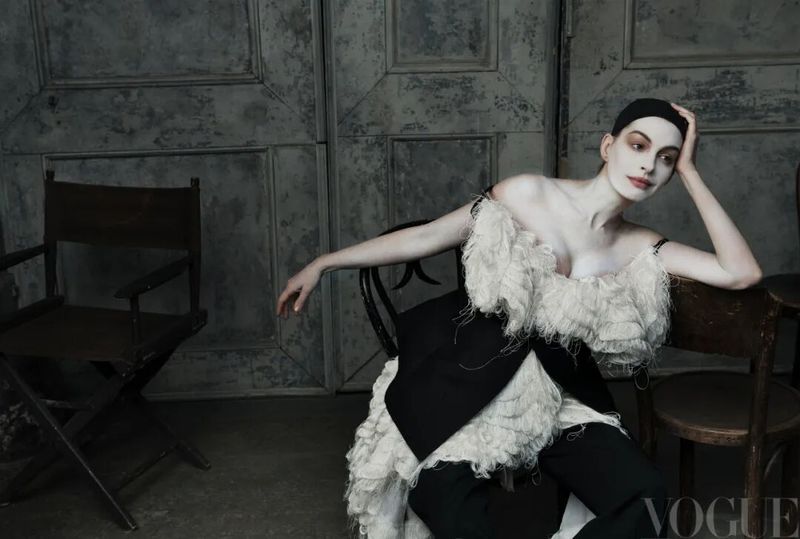 Anne Hathaway in theatrical white makeup and a ruffled cream top seated against a weathered panelled wall.