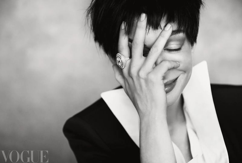 Black and white portrait of Anne Hathaway covering her smiling face with her hand, wearing a large ring and sharp-collared blazer.