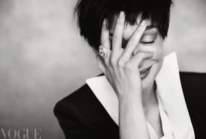 Black and white portrait of Anne Hathaway covering her smiling face with her hand, wearing a large ring and sharp-collared blazer.