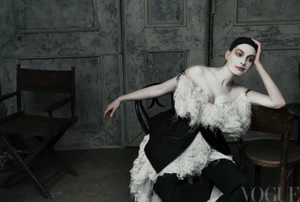 Anne Hathaway seated in a feathered cream dress and black trousers with dramatic white makeup against a distressed grey wall.