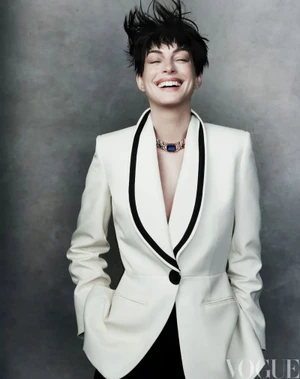 Anne Hathaway laughing in an off-white blazer with black shawl lapels and a gem choker necklace against a grey backdrop.