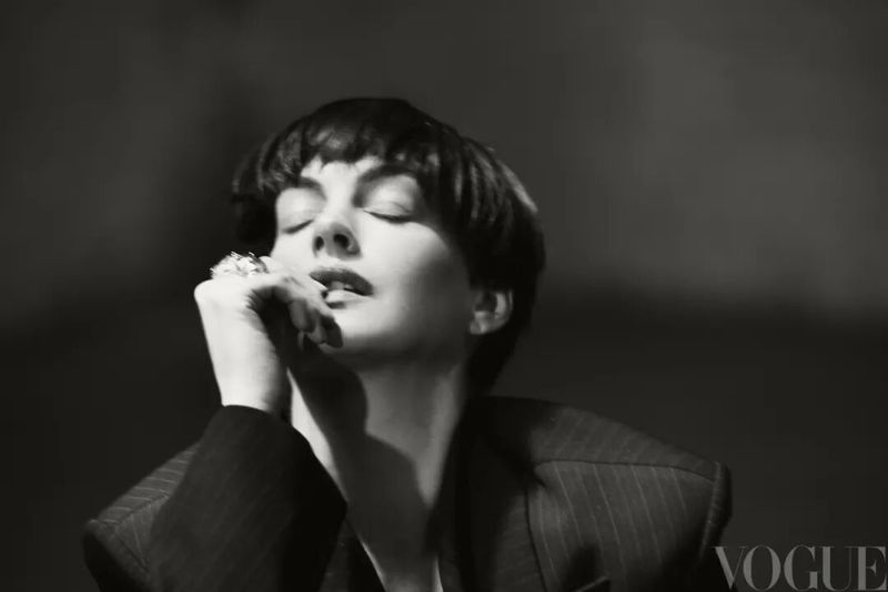 Anne Hathaway with a short pixie haircut and eyes closed posing in a dark pinstripe blazer with a statement ring.