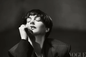 Anne Hathaway with a short pixie haircut and eyes closed posing in a dark pinstripe blazer with a statement ring.