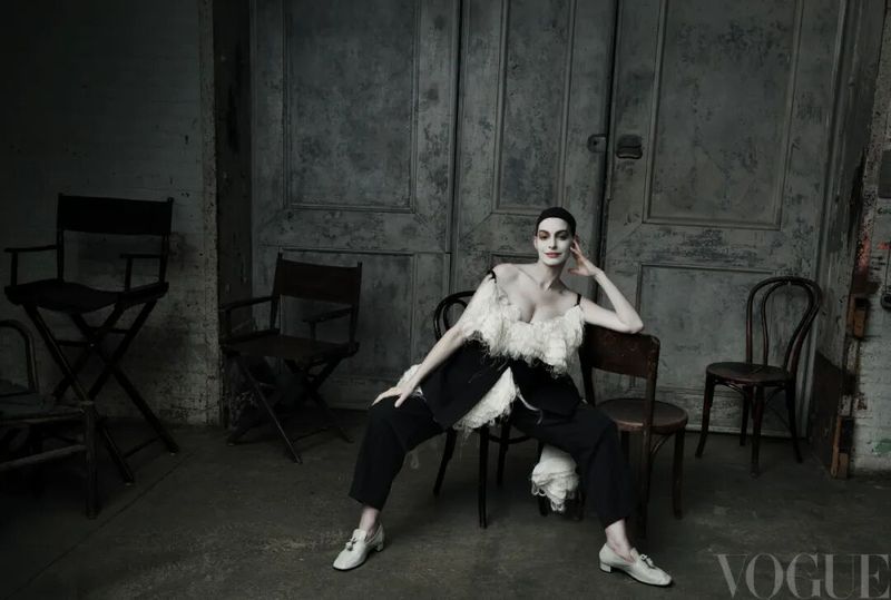 Anne Hathaway seated in a white ruffled top and black trousers surrounded by empty chairs for Vogue China.