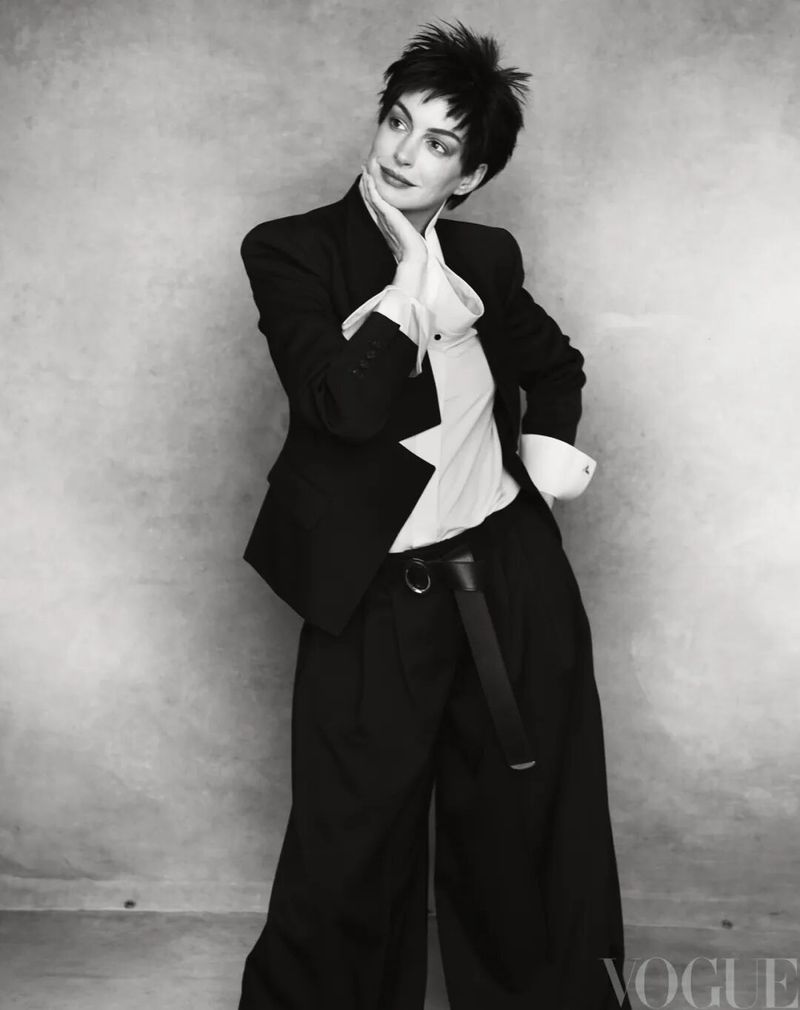 Anne Hathaway posing in an oversized black blazer, wide-leg trousers, and white shirt against a textured studio backdrop.