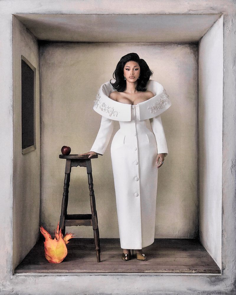 Cardi B standing in a sculptural white coat dress inside a painted alcove next to a dark wooden stool with an apple and a flame prop.