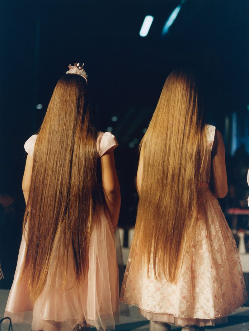Two young girls with long, straight, honey-blonde hair wear pink dresses as they stand indoors in an event space with dark background.
