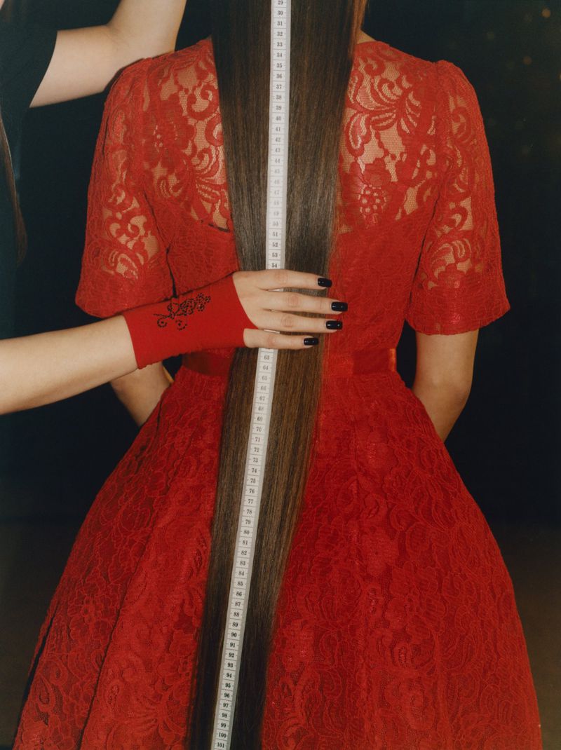 Back view of woman in red dress with extra-long straight hair being measured by hands with red sleeves and black manicure.