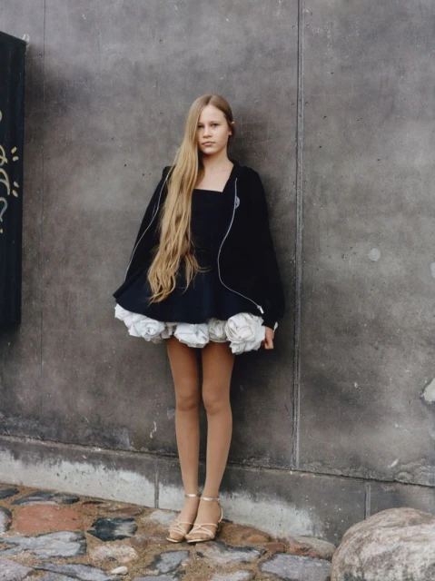 Young woman in a black dress with white flower detailing poses against a concrete wall, styled with long blonde hair and strappy heels.