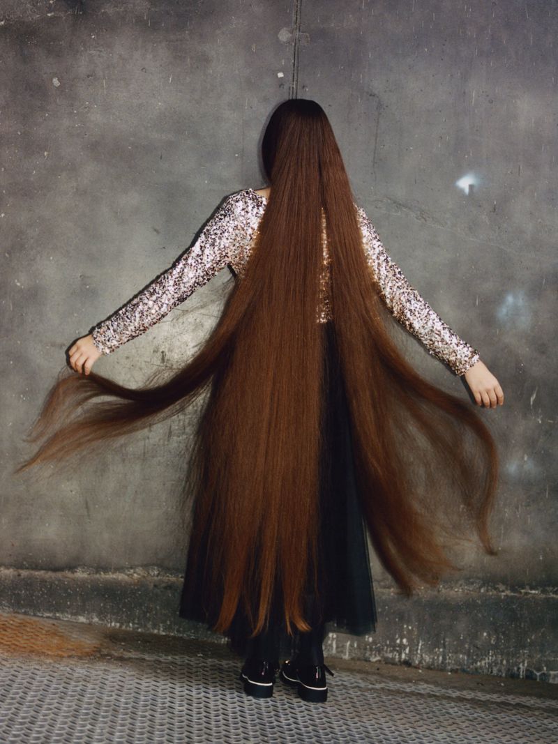 Back view of a person with long, flowing brown hair, wearing a sequined top and black skirt, standing against a textured gray wall.