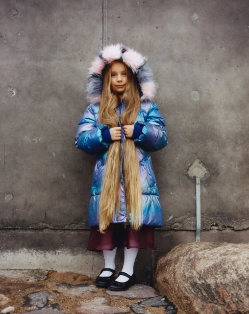 Girl wearing iridescent puffer coat with fur hood stands against a grey wall. Her long blonde hair is worn loose and straight.