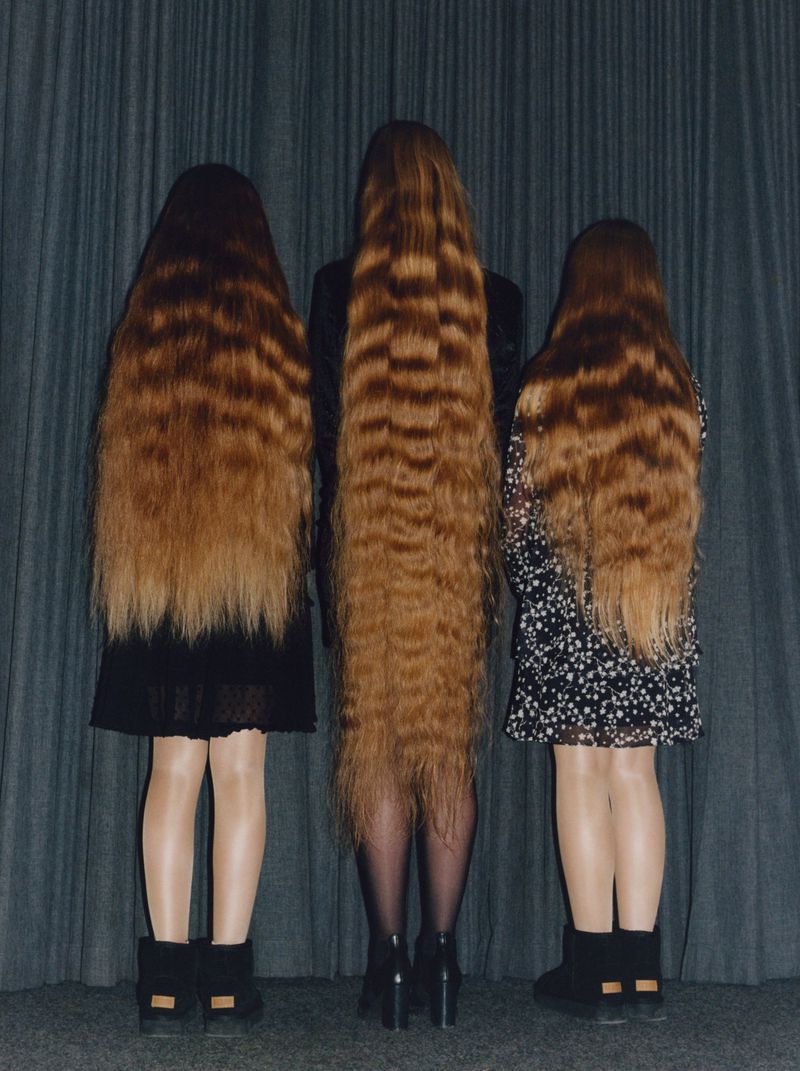 Three people with long brown crimped hair stand close together, their backs to the camera, wearing short dresses and dark footwear.
