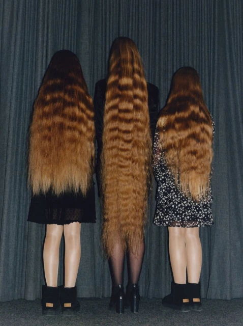 Three people with long brown crimped hair stand close together, their backs to the camera, wearing short dresses and dark footwear.