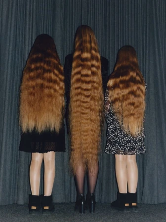 Three people with long brown crimped hair stand close together, their backs to the camera, wearing short dresses and dark footwear.