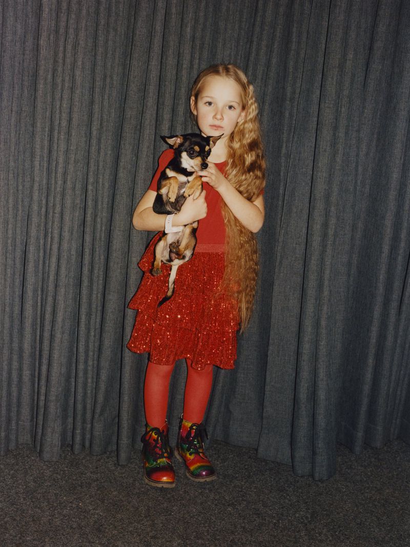 Little girl with long blonde hair wearing a red dress and holding a chihuahua-mix breed dog against a grey curtain backdrop.