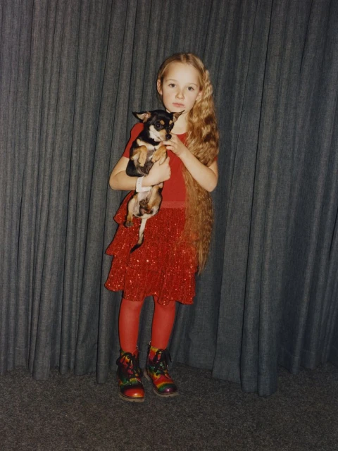 Little girl with long blonde hair wearing a red dress and holding a chihuahua-mix breed dog against a grey curtain backdrop.