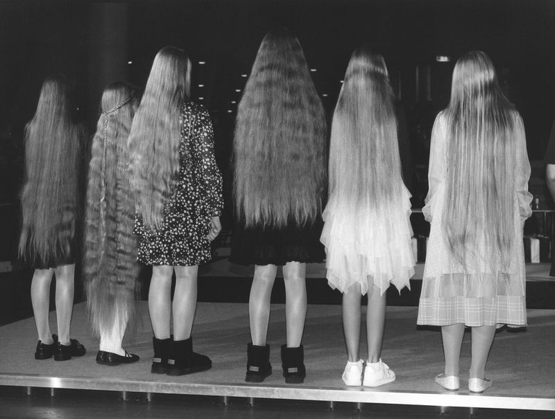 A grayscale image displays six young women from the rear, showcasing their long hair that reaches their lower backs, all standing on a platform.