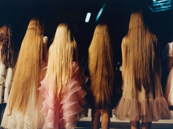 Back view of fashion models with long hair, wearing frilly dresses in a dimly lit indoor setting with a neutral color palette.