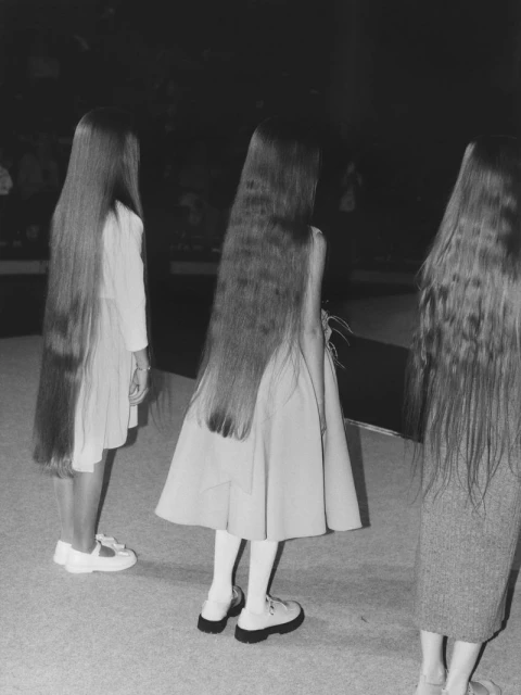 Three young girls stand with their backs to the camera, showcasing their long, dark hair and matching dresses in a dimly lit setting.