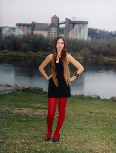 A full shot of a young woman in a black dress and red tights, hands on hips, standing in a grassy area near a river and industrial buildings.