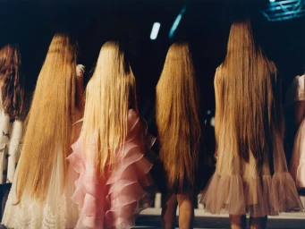 Group of young women with exceptionally long blonde hair seen from behind, wearing layered dresses.