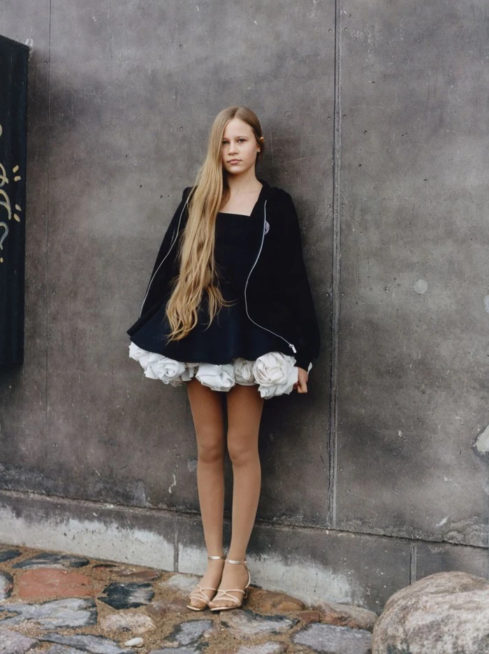 A young woman with very long blonde hair wearing a black jacket and a black dress with a ruffled white hem against a concrete wall.