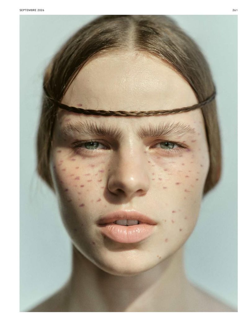 Close-up beauty portrait of a woman with a thin braid across her forehead, faux freckles, and feathered eyebrows on a pale backdrop.