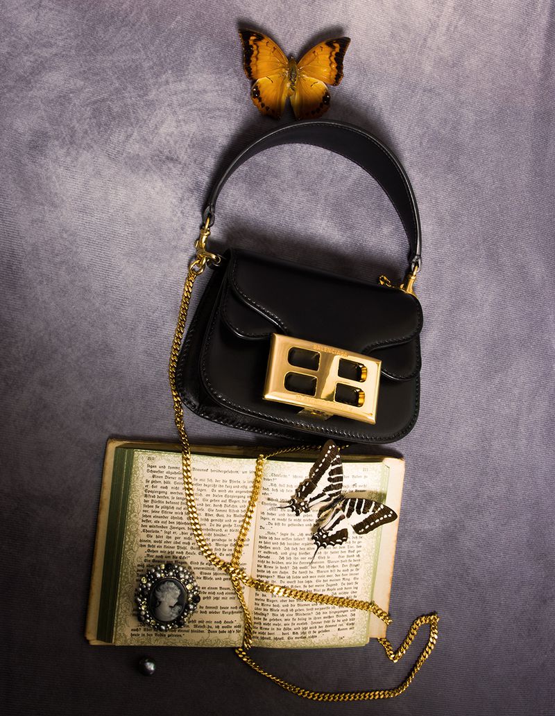 Black leather handbag with gold hardware displayed beside an open vintage book, cameo brooch, and two butterfly specimens.