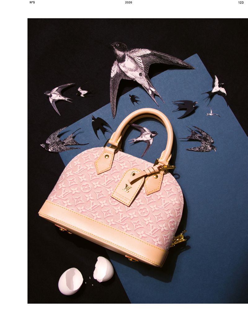 Pink monogram mini handbag surrounded by cutout paper swallows and broken eggshells on a blue and black surface.