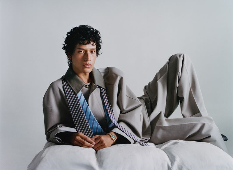 Model Magdaleno Delgado reclines in an oversized grey suit and draped striped ties against a plain studio backdrop.