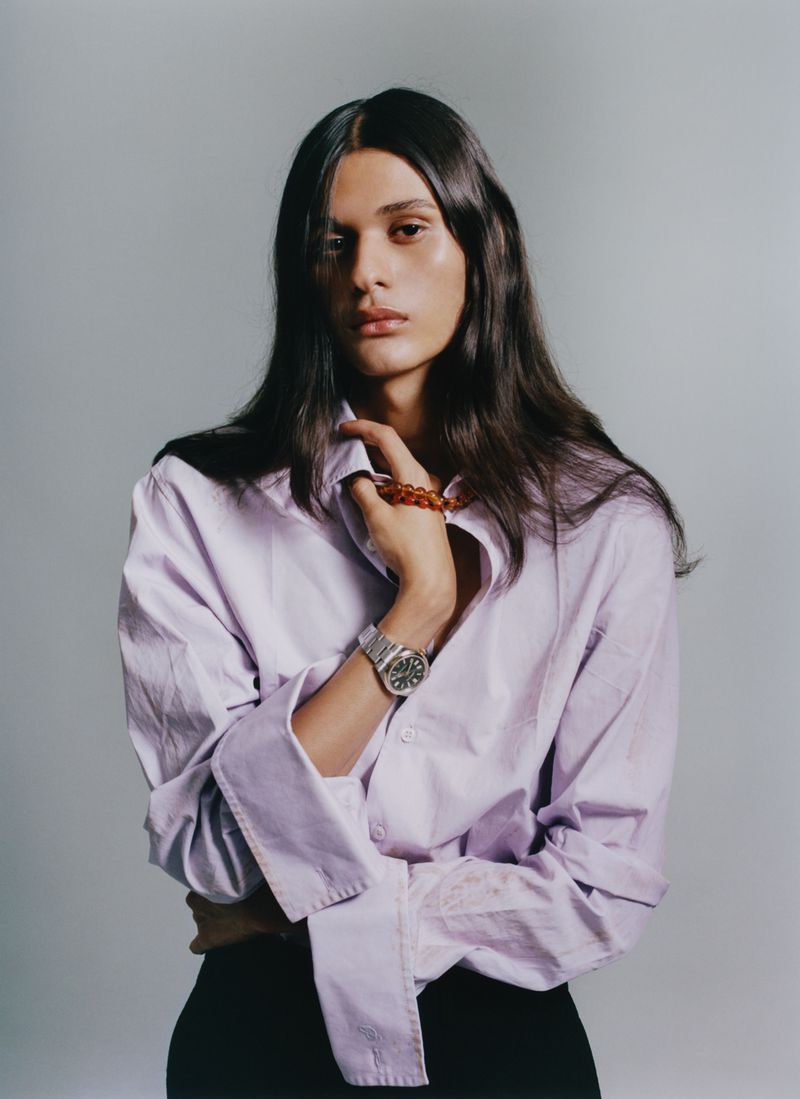 Model Magdaleno Delgado posing in an unbuttoned lilac dress shirt with a stainless steel wristwatch against a grey studio backdrop.