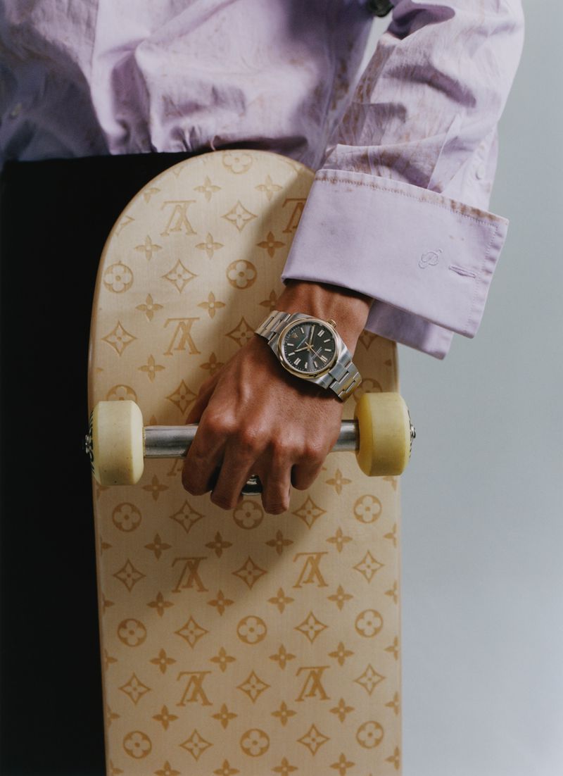 Close-up of a person in a lilac button-down shirt holding a monogram skateboard by its metal truck while wearing a steel wristwatch.