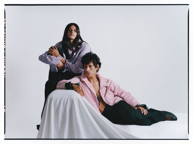Two fashion models posing against a white studio background with white draped fabric, wearing lilac and pink shirts.