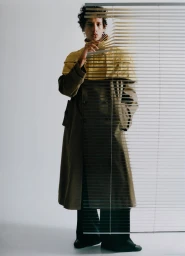 Model Magdaleno Delgado wearing a trench coat with a yellow overlay, peering through horizontal window blinds in a studio setting.