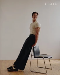 Man in a cream t-shirt and wide-leg black trousers leaning back against a black modern chair on a wooden studio floor.