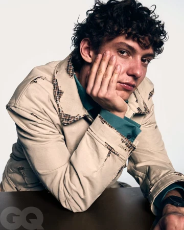 Kimi Antonelli rests his chin on his hand wearing a beige jacket over a teal shirt against a white studio background.