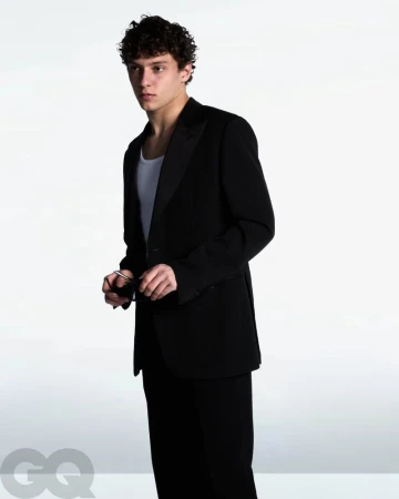 Andrea Kimi Antonelli wearing a black tailored blazer over a white tank top and holding sunglasses against a studio backdrop.