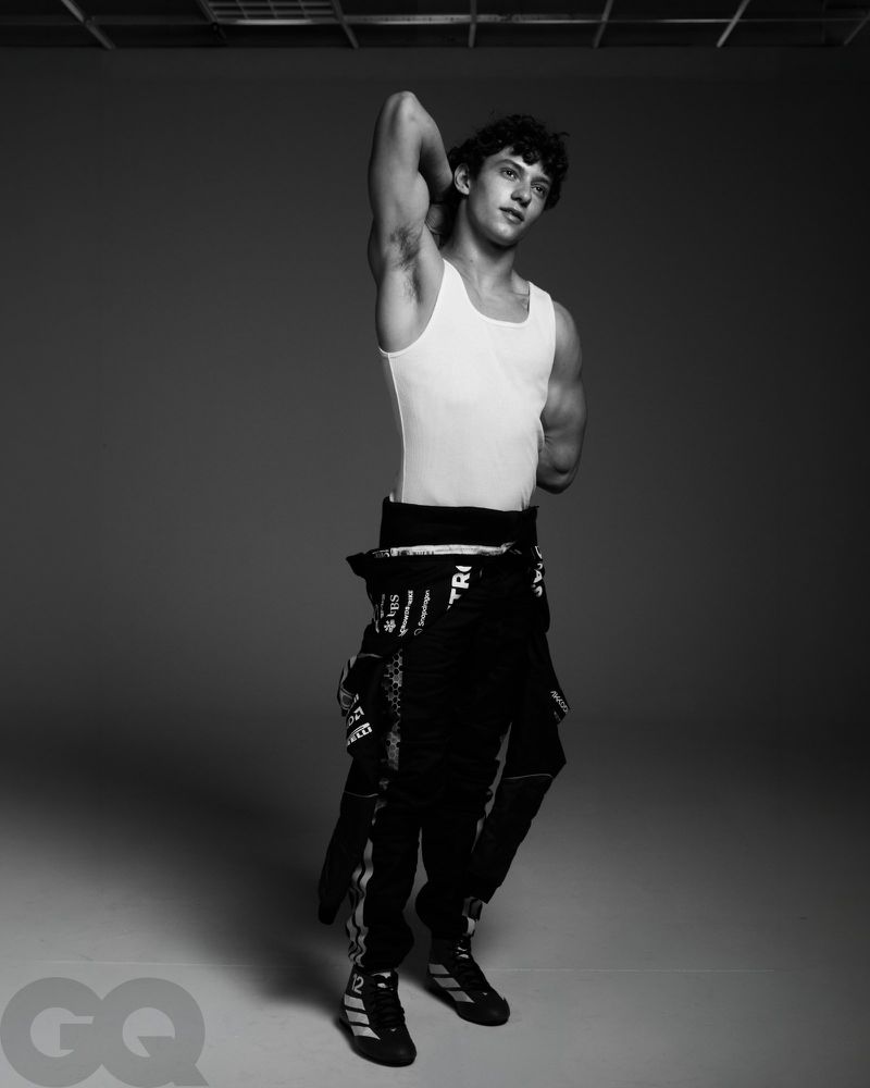 Black and white portrait of Kimi Antonelli in a white tank top and half-worn racing suit, posing against a grey studio background.