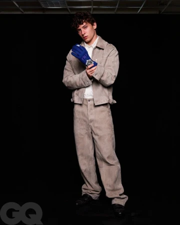 Model in a beige jacket and matching trousers wearing a blue racing glove and watch against a black studio background.