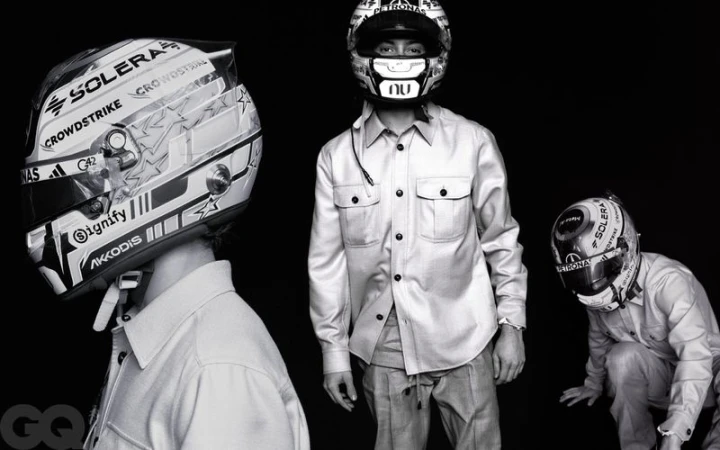 Monochrome editorial of three figures in light jackets wearing Mercedes AMG Petronas racing helmets against a black backdrop.