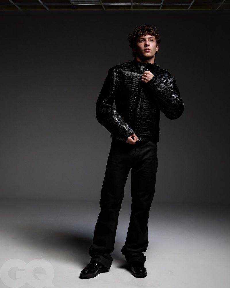 Young man standing in a black crocodile-embossed leather jacket and black trousers against a dark grey studio background.