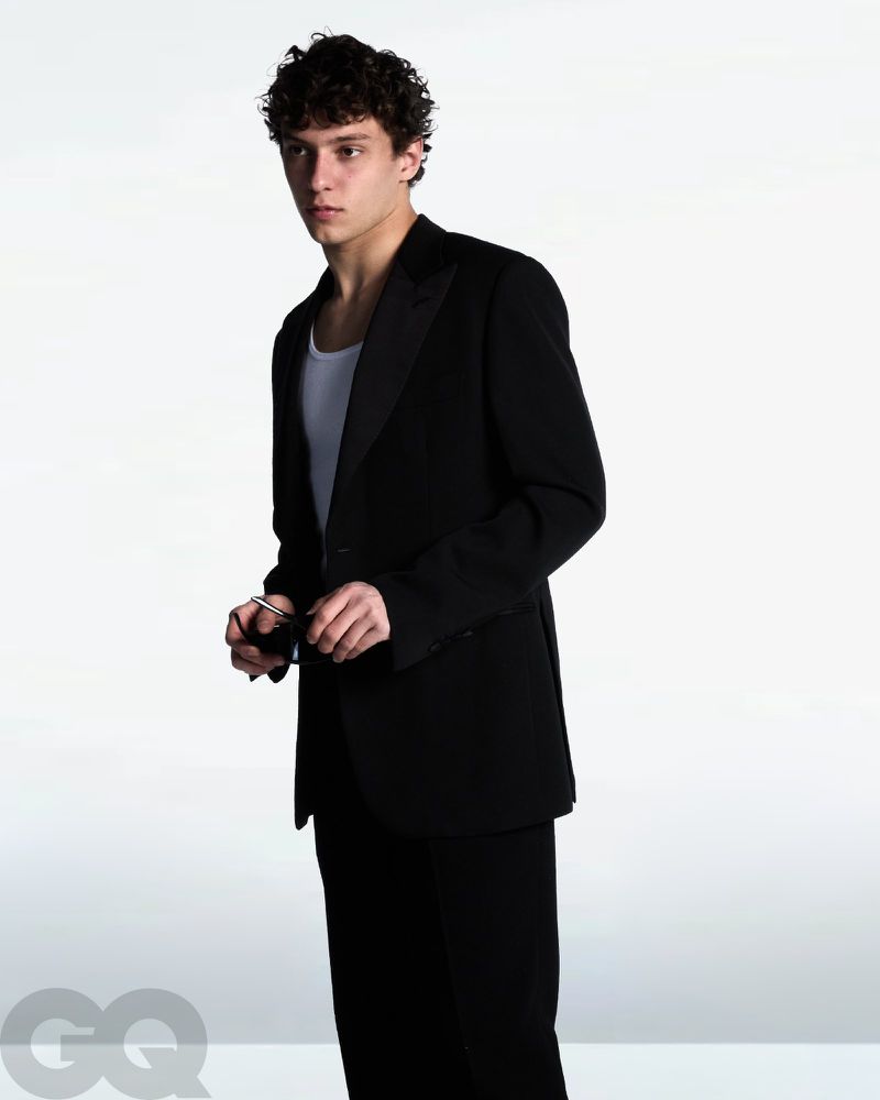 Kimi Antonelli wearing a black blazer over a white tank top while holding sunglasses against a white backdrop for GQ Italia.