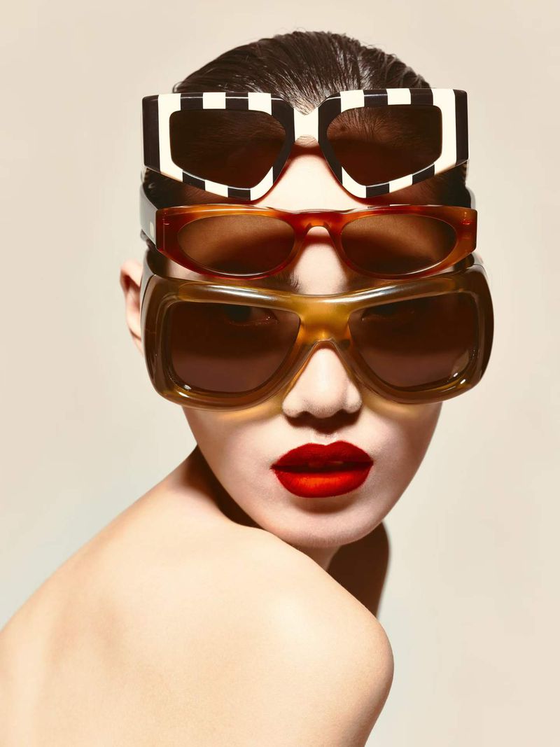 Close-up portrait of a model with slicked-back hair and bold red lips, wearing three pairs of stacked sunglasses, against a beige background.