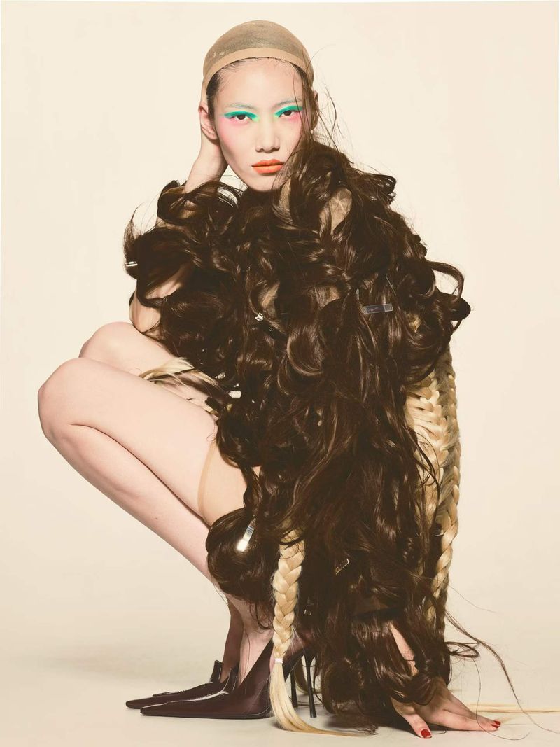 Model Hejia Li crouching draped in cascading brown hair extensions wearing a wig cap against a plain studio background.